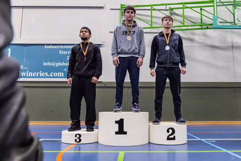 Three young wrestlers stand on a podium, wearing medals. The gold medalist stands tallest, center, looking composed.