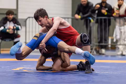 Young wrestler in red singlet drives opponent to the mat with intense focus during a competitive match.