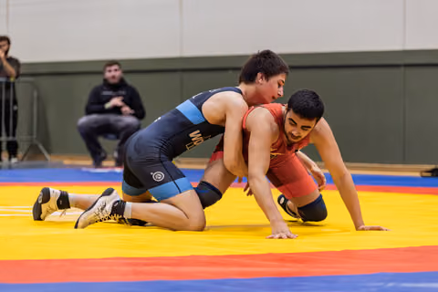 Young wrestler in blue singlet controls opponent in red from behind, both straining intensely on a colorful mat.