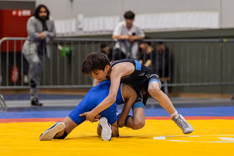 Two young wrestlers battle for control on a yellow mat, one in blue executing a takedown while spectators watch.