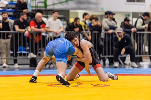 Two young wrestlers lock up on a yellow mat, one in blue singlet crouching low, the other in black staying balanced.
