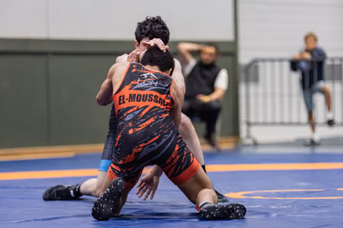 Wrestler El-Moussaed in orange singlet drives opponent to the mat in an intense grappling exchange.