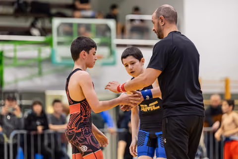Adult coach facilitates handshake between two young wrestlers in singlets, overseeing sportsmanship on the mat.