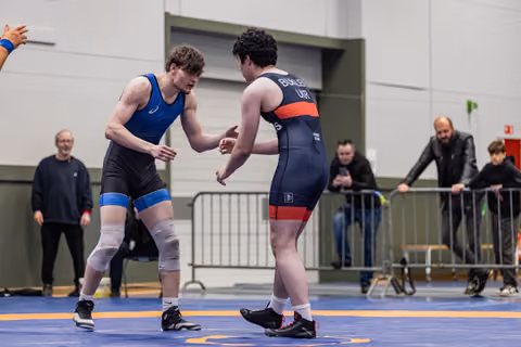 Two young male wrestlers reach toward each other in a pre-match handshake on a blue mat, spectators watching behind barriers.