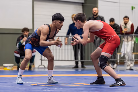 Two teen wrestlers in intense face-off stance on blue mat, crowds watching behind barrier.