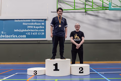 Young man smiling proudly on 1st place podium wearing gold medal, older man standing beside 2nd place podium in wrestling shirt.