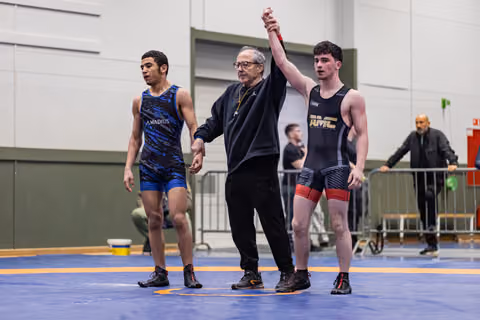 Referee raises the right arm of a victorious wrestler in black singlet, while defeated opponent in blue looks down.