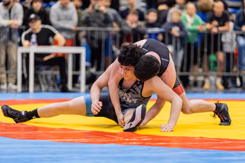 Young wrestler in black singlet resists opponent's takedown attempt, legs spread wide on a yellow and red mat.