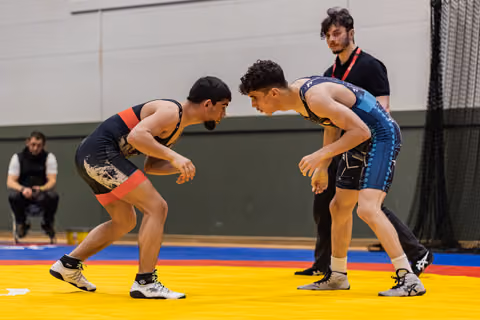 Two young wrestlers in a tense standoff, leaning toward each other on a yellow mat, referee watching closely behind.
