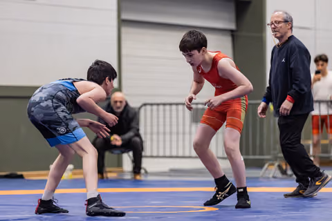 Two youth wrestlers in blue and red singlets crouch in tense standoff on blue mat, referee watching nearby.