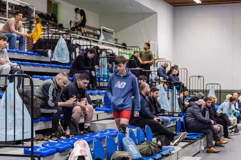 Teen in New Balance hoodie and red shorts walks pensively through blue stadium seats among waiting athletes.