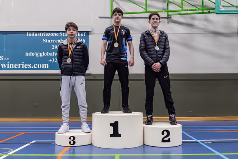 Three young male wrestlers stand on a podium, wearing bronze, gold, and silver medals, with solemn expressions in an indoor sports hall.