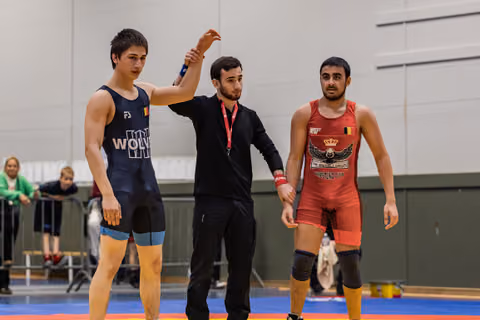 Referee raises the arm of a victorious wrestler in blue, while the defeated opponent in red stands nearby.