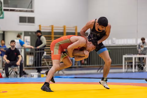 Wrestler in black singlet aggressively controls bent-over opponent in red during indoor match on yellow mat.