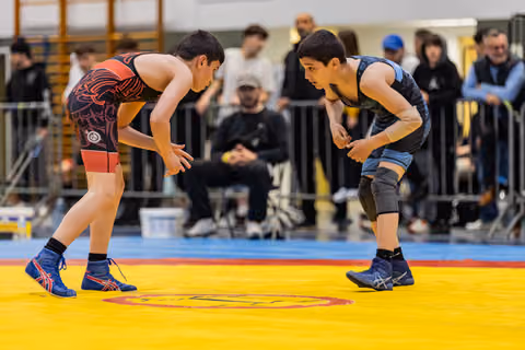 Two young wrestlers crouch in defensive stances, locked in intense eye contact before engaging on a yellow competition mat.
