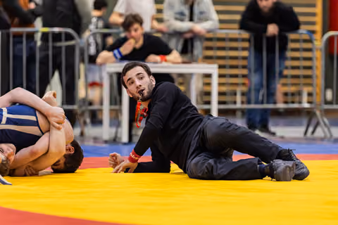 Referee lying on yellow mat blows whistle while closely watching two wrestlers grapple during indoor tournament.