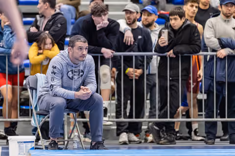 Coach in Under Armour hoodie sits intently at ringside, hands clasped, watching the match with focused concentration.