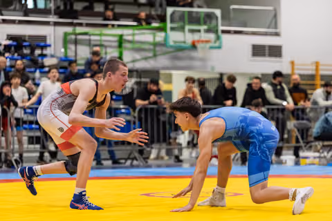 Two young wrestlers in intense standoff; one crouching low in blue, the other upright in white, crowd watching.