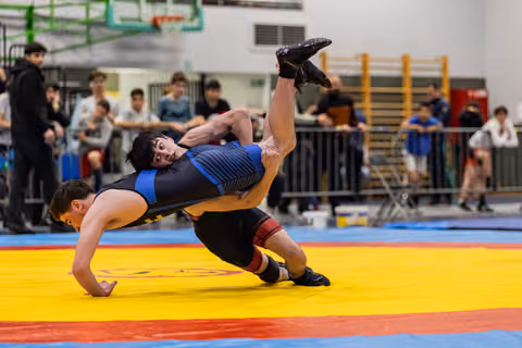 Young wrestler in blue singlet performs a powerful suplex throw, launching opponent airborne on colorful competition mat.