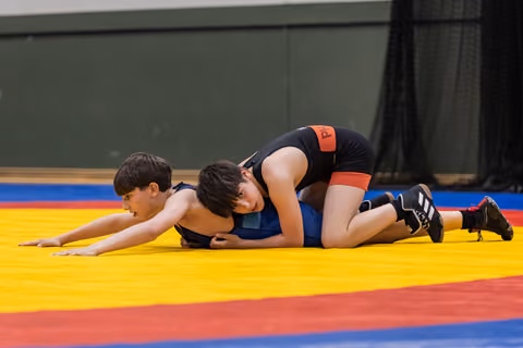 Two young wrestlers on a yellow mat, one in black pinning the other in blue, both showing intense focus and effort.