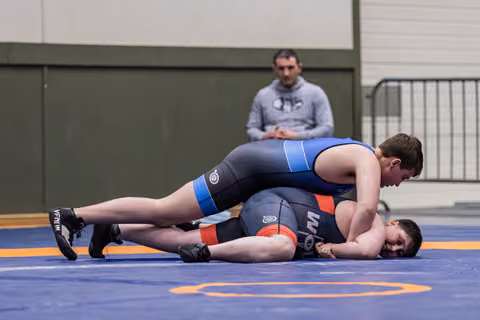Young wrestler in blue singlet pins opponent to the mat while a coach watches intently from the background.
