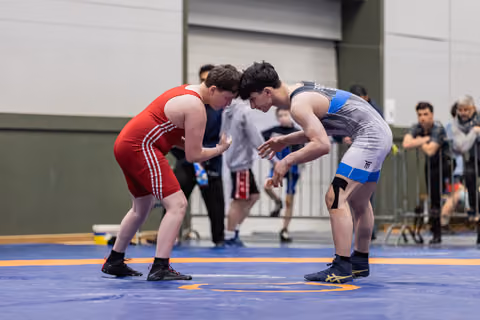 Two young wrestlers crouch face-to-face in a tense standoff on a blue mat, spectators watching in the background.