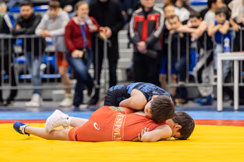 Wrestler in red singlet pinned to the mat by blue opponent, crowd watching intently in the background.