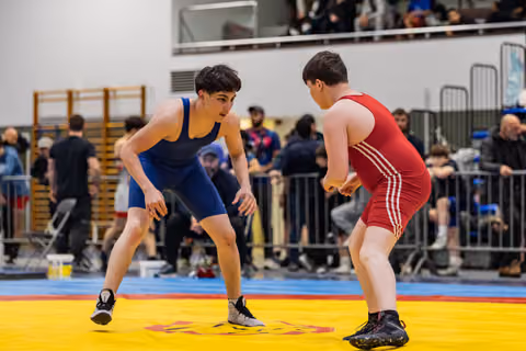 Two young wrestlers in blue and red singlets crouch in tense standoff on colorful mat, crowd watching behind barriers.