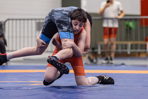 Young wrestler in red singlet aggressively grips opponent's leg, attempting a takedown on a blue competition mat.
