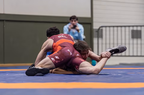 Belgian wrestler in red singlet controls opponent on the mat during an intense grappling match indoors.