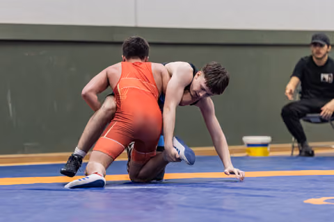 Teen wrestler in black singlet drives forward aggressively while opponent in red struggles on knees; referee observes from sideline.