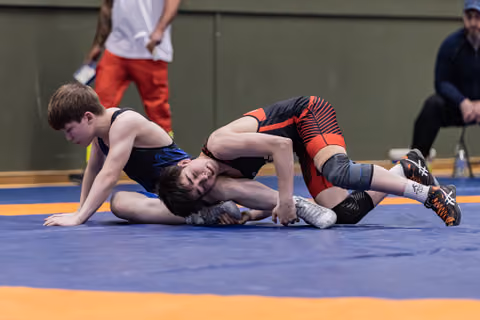 Two youth wrestlers compete on a blue mat, one pinning the other down with focused intensity during a match.