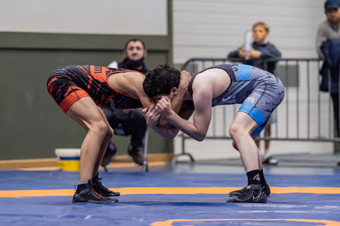 Two wrestlers locked in an intense clinch, bent low in a defensive stance on a blue competition mat.