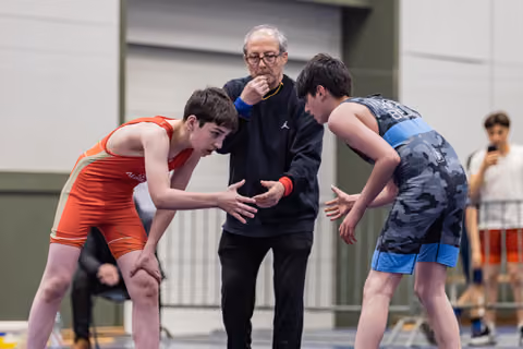 Two young wrestlers lean in to shake hands before a bout, overseen by a referee blowing a whistle indoors.
