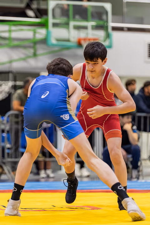 Two teen wrestlers in red and blue singlets square off on a yellow mat, the red wrestler focused and alert.