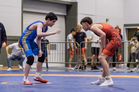 Two teen wrestlers in blue and red singlets crouch low, locked in intense eye contact before their match on a blue mat.