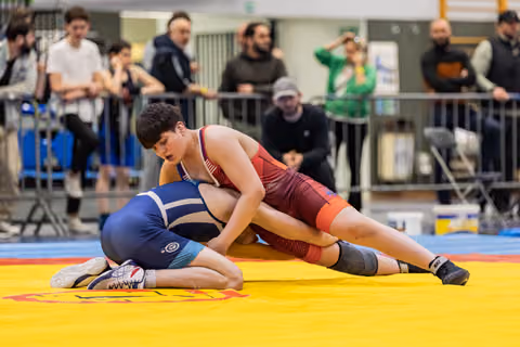 Young wrestler in red singlet dominates opponent in blue, pinning them down on yellow competition mat indoors.