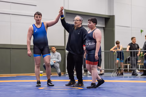 Referee raises the hand of a young wrestler in blue singlet declaring victory, as opponent stands nearby on blue mat.