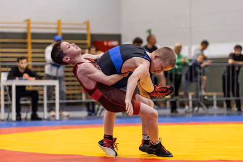 Young wrestler in red singlet hoists opponent off the mat during an intense match in a gymnasium.