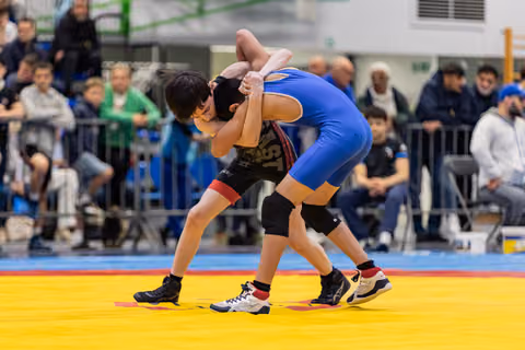 Two young wrestlers locked in a grappling hold on a yellow mat, crowds watching in a busy sports hall.