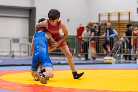 Young wrestler in red singlet controls kneeling opponent in blue during intense youth wrestling bout on colorful mat.