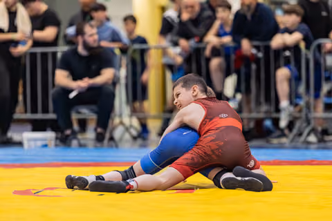 Two young wrestlers battle on a yellow mat, one in red pinning the other in blue, faces tense with effort.