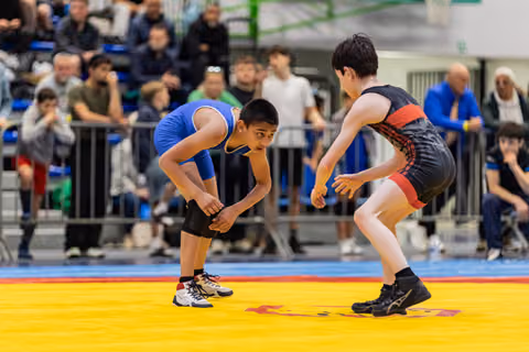 Two boys in wrestling stance face each other intently on a yellow mat, crowd watching in the background.