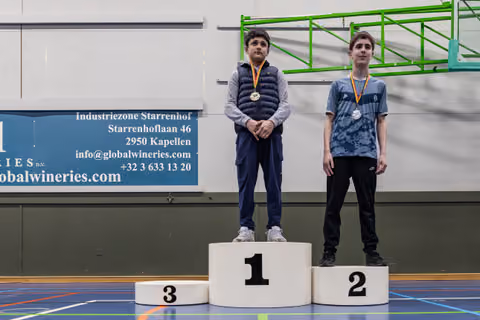 Two young boys stand on a podium, first and third place winner with gold medal, second place with silver, indoors.