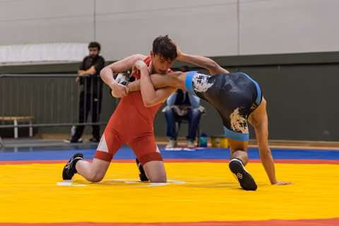 Young wrestler in red singlet on one knee, gripping opponent in dark blue singlet in intense clinch on yellow mat.