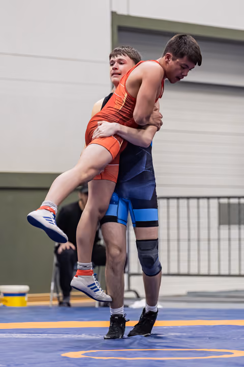 Young wrestler in blue lifts opponent in red off the mat, showing strength and determination during a match.