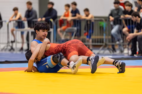 Teen wrestler in red singlet pins opponent to the mat, focused expression, surrounded by watching crowd.