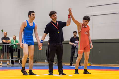 Referee raises the red-singlet wrestler's arm in victory while blue-singlet opponent stands defeated on yellow mat.