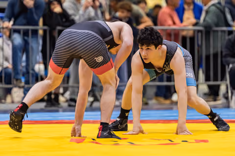 Two young wrestlers crouch in a tense standoff on a yellow mat, the facing athlete showing focused concentration.