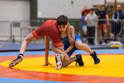 Two young wrestlers battle intensely on a yellow mat, one in red lifting his opponent in blue during a takedown attempt.